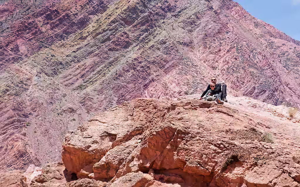 Touristen sitzen entspannt auf einem roten Felsvorsprung und genießt die Aussicht auf den Cerro de los Siete Colores in Purmamarca, Jujuy, Argentinien. Im Hintergrund erstrecken sich die gewaltigen, diagonal verlaufenden Sedimentschichten der Anden in lebendigen Rosa-, Rot- und Ockertönen unter strahlender Sonne.